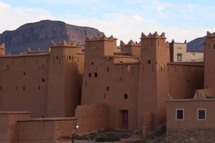 Mudbrick kasbah in N'Kob showing tall towers, crenellations, and rugged terrain behind.