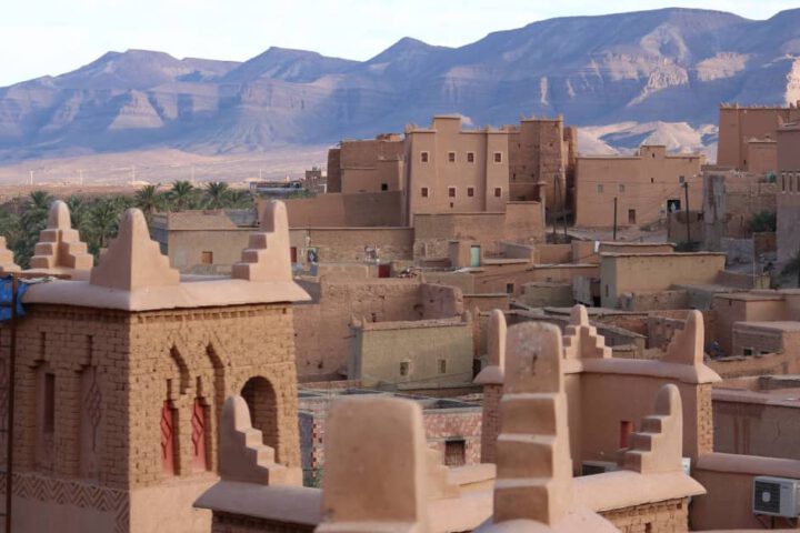 N'Kob village earthen houses with Jbel Saghro mountains and a palm grove under a clear sky.
