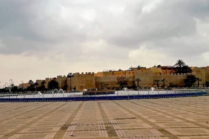 Wide paved area of 20 August Place (Sahat 20 Ghoucht) in Taroudant with historic crenelated city walls and palm trees in the background.