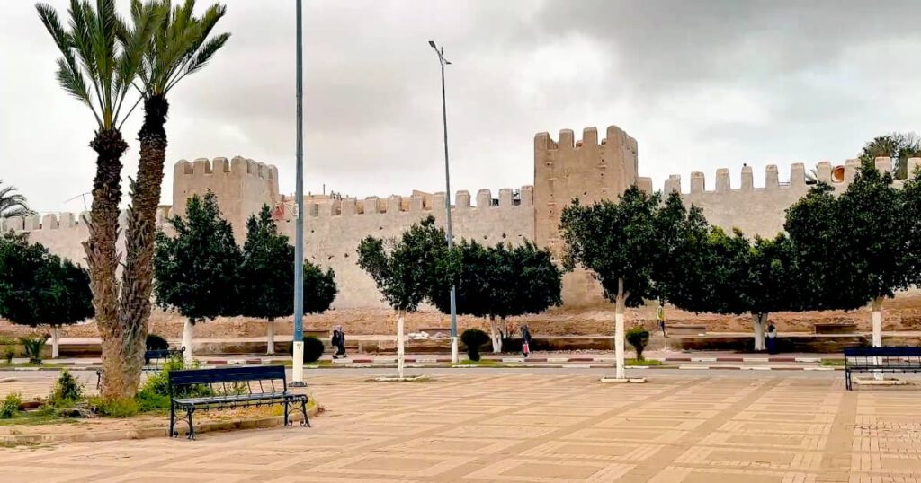 Public Square and Historic Ramparts of Taroudant, Morocco Wide paved public square with park benches and trees in front of the historic ocher-colored Taroudant city walls under a cloudy sky.