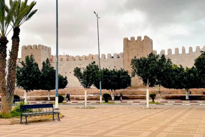 Wide paved public square with park benches and trees in front of the historic ocher-colored Taroudant city walls under a cloudy sky.