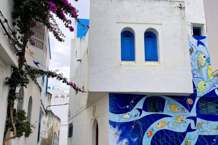 Narrow whitewashed alley in Asilah featuring a blue fish mural and blooming bougainvillea.