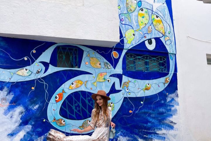 Traveler standing in front of a large blue fish mural on a white wall in Asilah.