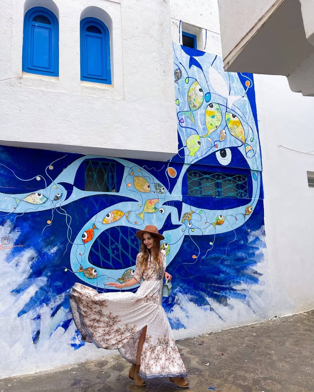 Traveler standing in front of a large blue fish mural on a white wall in Asilah.