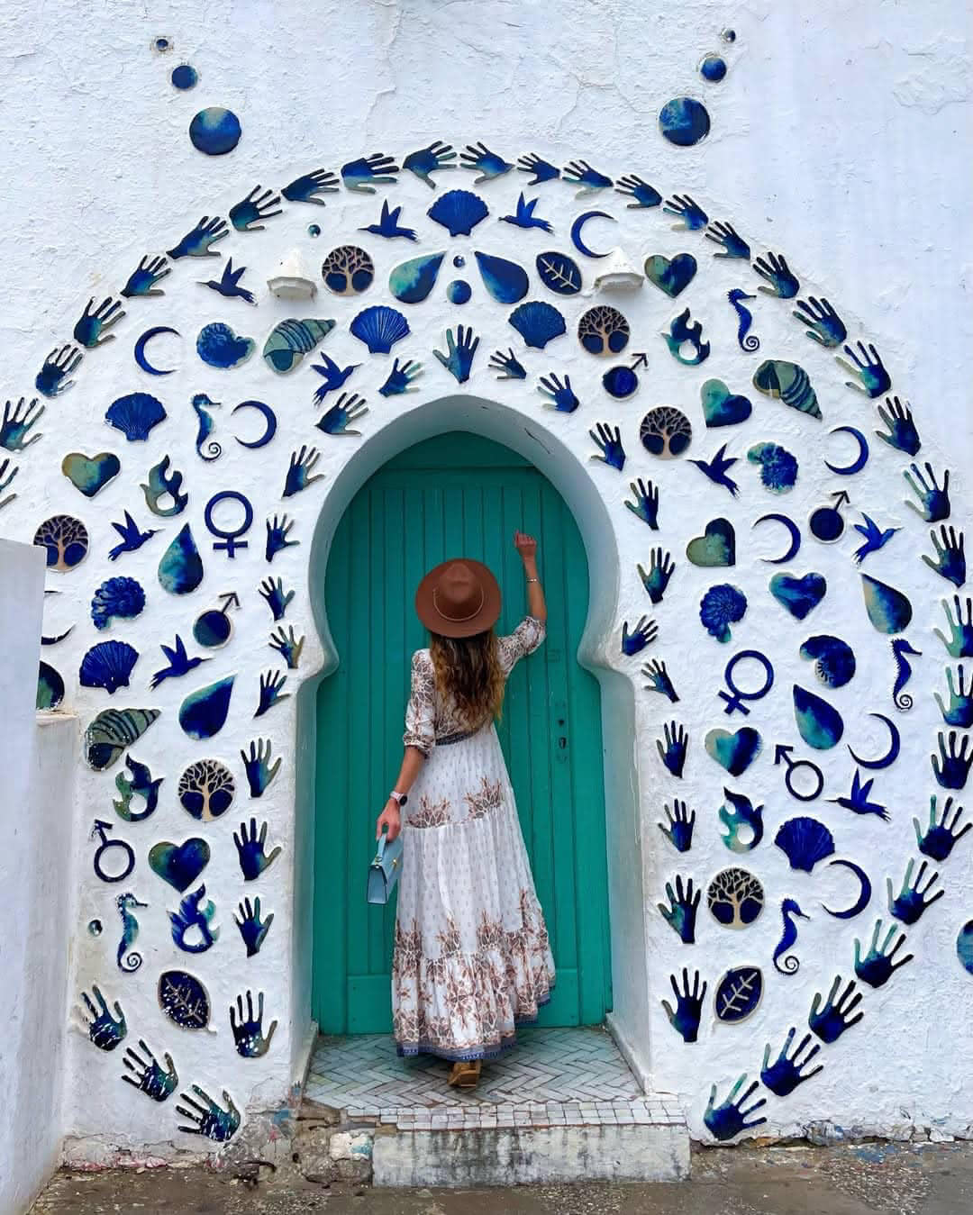 Traveler knocking on a turquoise arched door surrounded by a blue mosaic of symbols in Asilah.