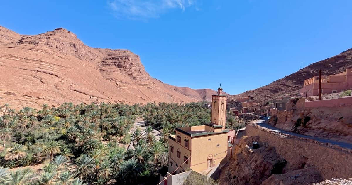 Wide panoramic view of a lush green palm grove and winding stream at the base of steep, rugged red rock canyon walls in Ait Mansour Gorge, Morocco.