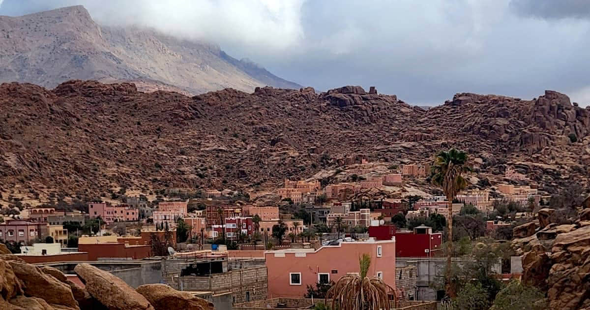 A Wide panoramic view of Tafraout village with colorful pink and red buildings at the base of rugged granite mountain slopes under a cloudy sky in Morocco.
